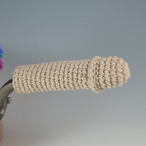 May include: A beige crocheted pot handle cover. The cover is crocheted in a simple stitch and has a slight twist at the end. The handle cover is designed to protect hands from hot pots.