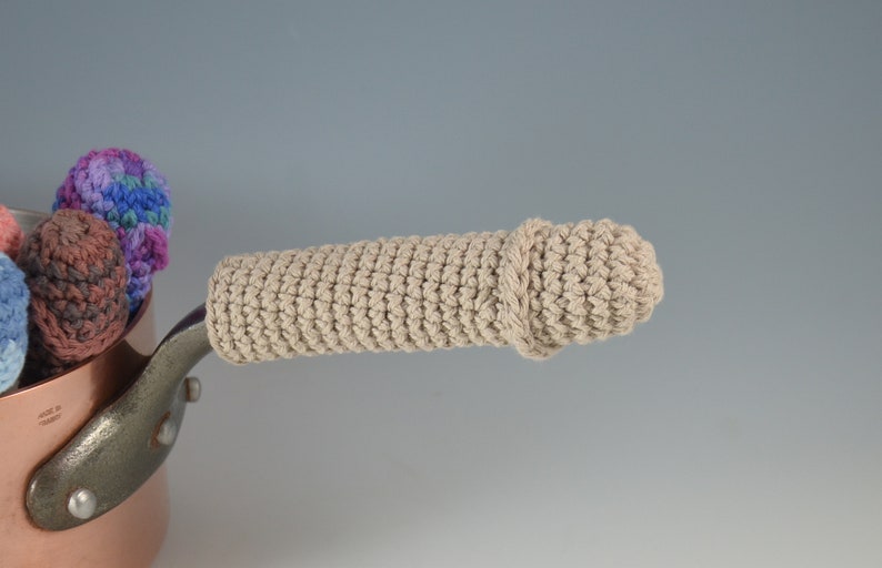 May include: A beige crocheted pot handle cover. The cover is crocheted in a simple stitch and has a slight twist at the end. The handle cover is designed to protect hands from hot pots.