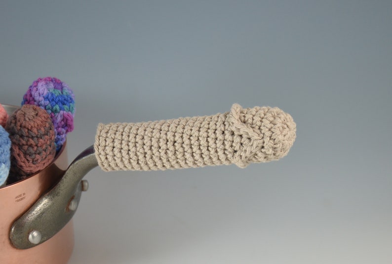 May include: A beige crocheted pot handle cover with a rounded end. The handle cover is on a copper pot.