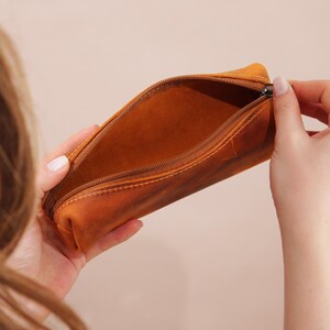 May include: Open, brown leather pencil case with a zipper closure. The pencil case is held open, revealing the interior. The leather has a distressed appearance with visible stitching.