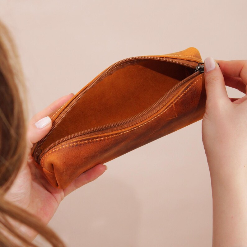May include: Open, brown leather pencil case with a zipper closure. The pencil case is held open, revealing the interior. The leather has a distressed appearance with visible stitching.