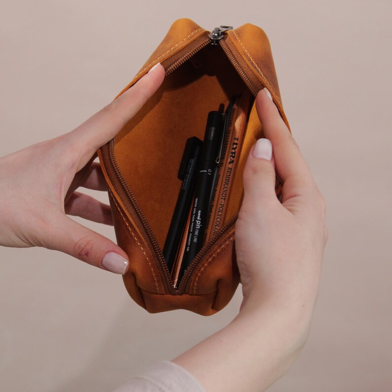 May include: A tan leather pencil case, partially unzipped, revealing black pens and a colored pencil. The case has a zipper closure and is held open by hands. The interior is a lighter shade of tan.