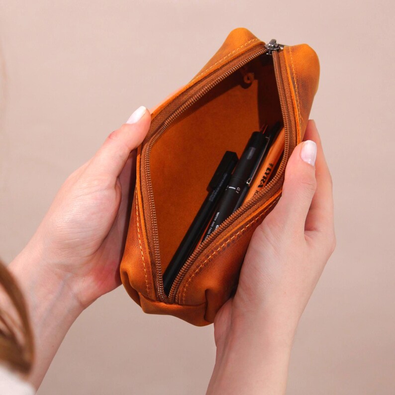 May include: Open, orange-brown leather pencil case with a zipper, revealing black pens inside. The case is held open by hands, showcasing the interior and the pens. The leather has a slightly distressed appearance.
