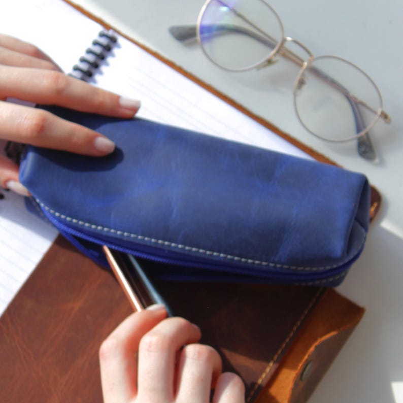 May include: A blue leather pencil case with a zipper, white stitching, and a pen inside. The case is on top of a notebook and a brown leather journal. Round eyeglasses rest on the notebook.