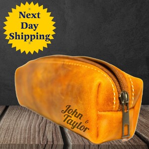 May include: A rectangular, tan leather pencil case with a bronze zipper and white stitching. The words "John & Taylor" are printed on the side. The pencil case is on a wooden surface.