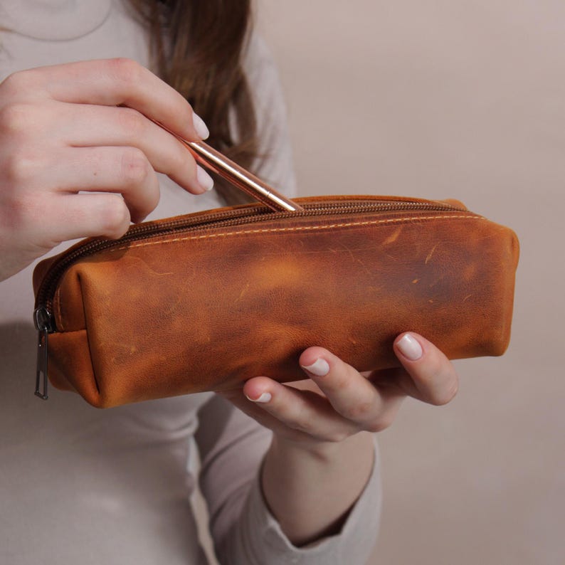 May include: A brown leather pencil case with a zipper closure. A rose gold pen is being placed inside the case. The pencil case has a distressed finish and is rectangular in shape. The case is held by a person.