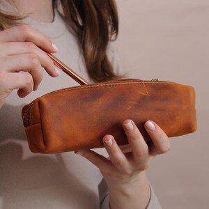 May include: A brown leather pencil case with a zipper closure. A rose gold pen is being placed inside the case. The pencil case has a distressed finish, giving it a vintage look. The case is rectangular and appears to be empty.