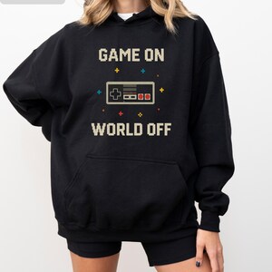 May include: Black hoodie with a pixelated video game controller graphic. The text "GAME ON WORLD OFF" is printed above and below the controller. The hoodie has a front pocket and a hood.
