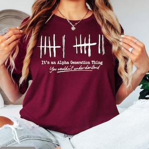May include: Maroon t-shirt with white text that reads "It's an Alpha Generation Thing, You wouldn't understand." The design includes vertical lines. The shirt is a crew neck.