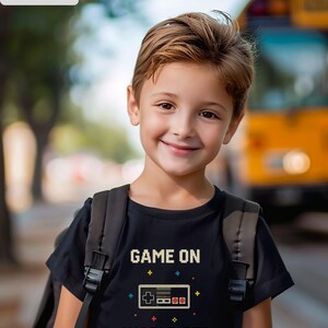 May include: A black t-shirt with the text "GAME ON WORLD OFF" and a pixelated video game controller design. The shirt is worn by a child with a backpack, smiling in front of a blurred background.