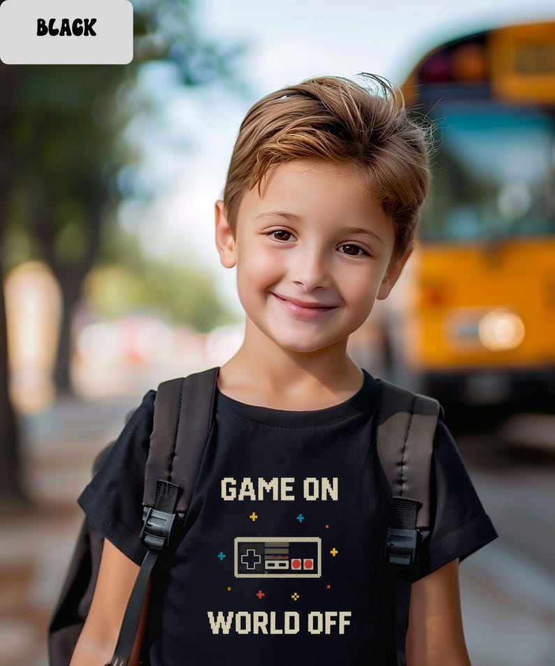 May include: A black t-shirt with the text "GAME ON WORLD OFF" and a pixelated video game controller design. The shirt is worn by a child with a backpack, smiling in front of a blurred background.