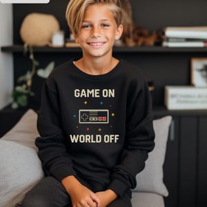 May include: Black sweatshirt with the text "GAME ON WORLD OFF" and a pixelated video game controller graphic. The sweatshirt is worn by a person.
