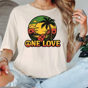 May include: Cream-colored t-shirt featuring a colorful graphic of a sunset, palm tree, and guitar within a circle. The words "ONE LOVE" are printed in bold, orange and yellow letters. The shirt is made of a soft material.