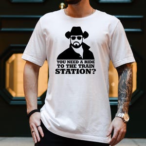 May include: White t-shirt featuring a black graphic of a man in a cowboy hat and sunglasses, with the text "YOU NEED A RIDE TO THE TRAIN STATION?". The shirt is a classic crew neck style.