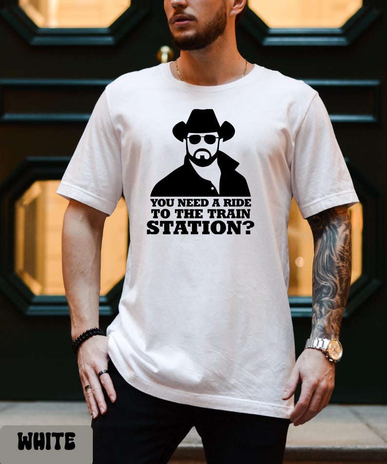 K&ouml;nnte beinhalten: Wei&szlig;es T-Shirt mit einer schwarzen Grafik eines Mannes mit Cowboyhut und Sonnenbrille und dem Text "YOU NEED A RIDE TO THE TRAIN STATION?". Das T-Shirt hat einen Rundhalsausschnitt.