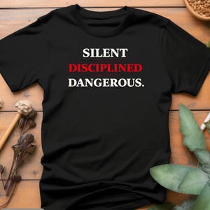 May include: Black t-shirt with the words "SILENT DISCIPLINED DANGEROUS." in white and red lettering. The shirt is displayed on a wooden surface with decorative elements.