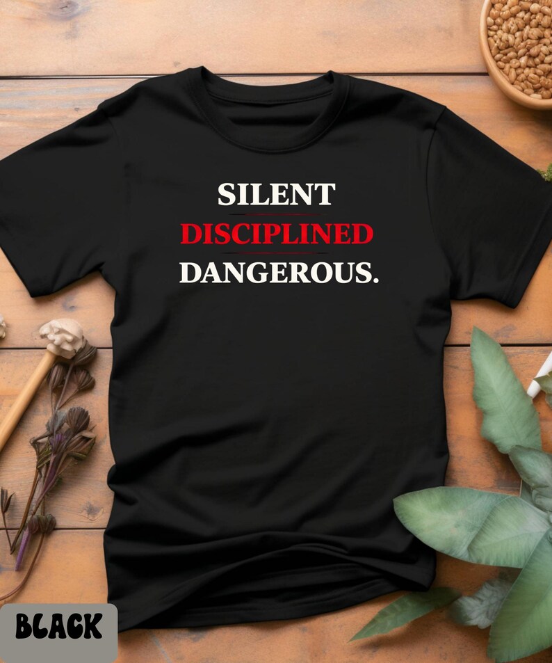 May include: Black t-shirt with the words "SILENT DISCIPLINED DANGEROUS." in white and red lettering. The shirt is displayed on a wooden surface with decorative elements.