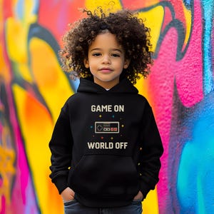 May include: A black hoodie with the text "GAME ON WORLD OFF" in white. The design includes a pixelated game controller. The child is wearing blue jeans and standing in front of a colorful graffiti wall.