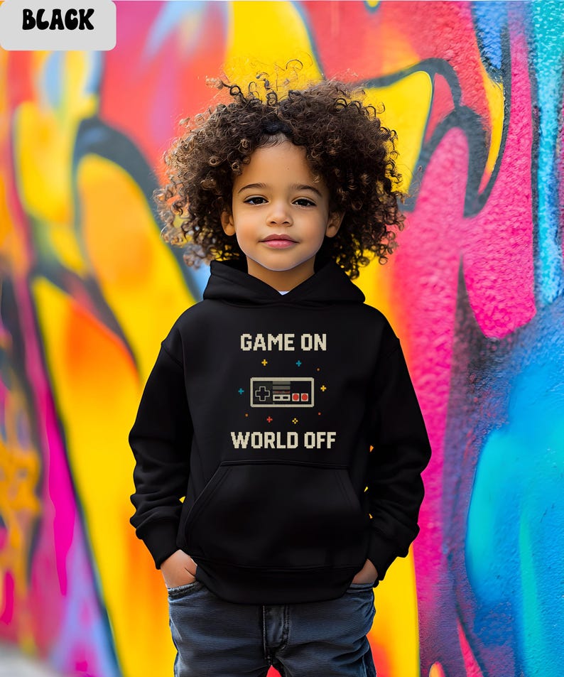 May include: A black hoodie with the text "GAME ON WORLD OFF" in white. The design includes a pixelated game controller. The child is wearing blue jeans and standing in front of a colorful graffiti wall.