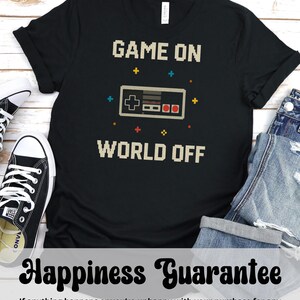 May include: Black t-shirt with the text "GAME ON WORLD OFF" in pixelated font, featuring a pixelated video game controller. The shirt is paired with black and white sneakers and blue jeans. The shirt also has a "Happiness Guarantee" message.