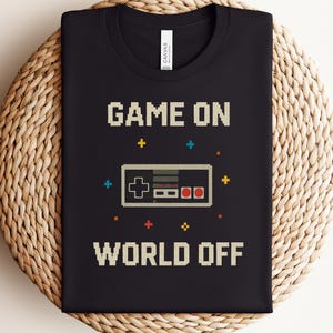 May include: Black t-shirt with the text "GAME ON WORLD OFF" in pixelated font. A pixelated controller graphic is centered below "GAME ON". The shirt is folded on a woven surface. The word "BLACK" is in a gray box at the bottom.