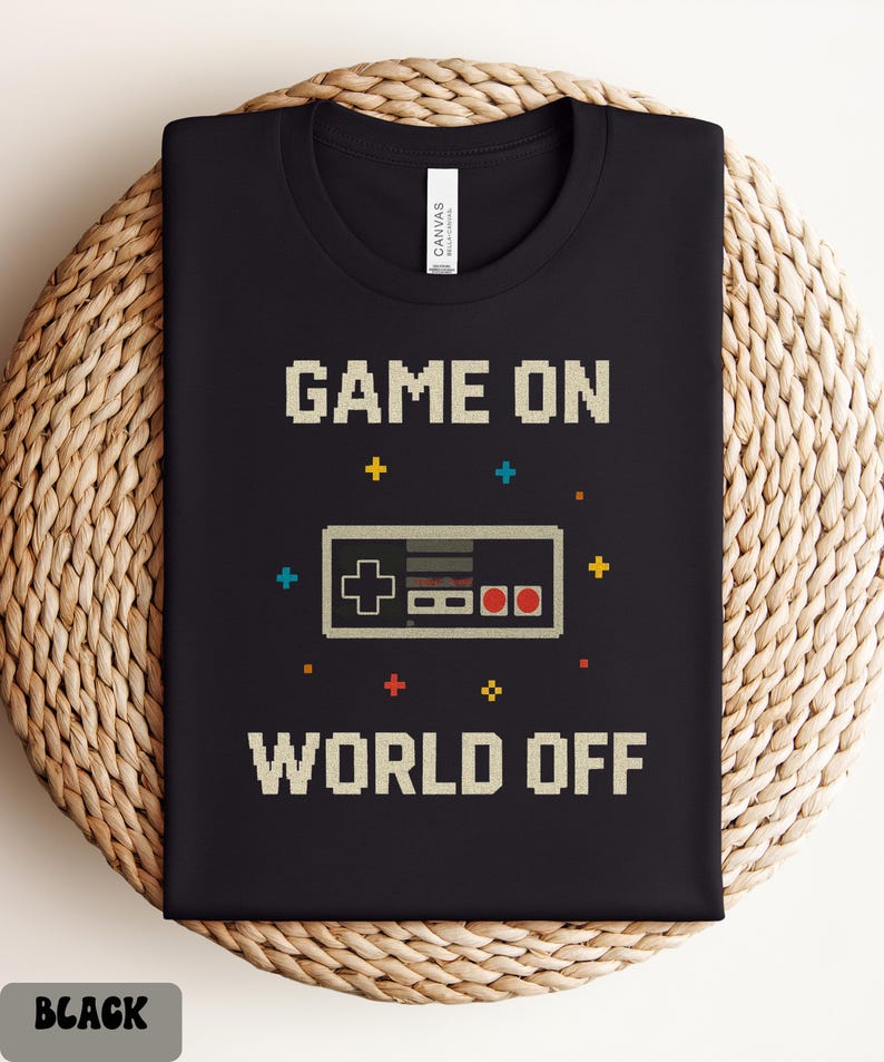 May include: Black t-shirt with the text "GAME ON WORLD OFF" in pixelated font. A pixelated controller graphic is centered below "GAME ON". The shirt is folded on a woven surface. The word "BLACK" is in a gray box at the bottom.