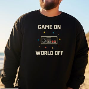 May include: Black sweatshirt with the text "GAME ON WORLD OFF" in pixelated white letters. A pixelated controller graphic is centered between the text. The sweatshirt is a crew neck style.