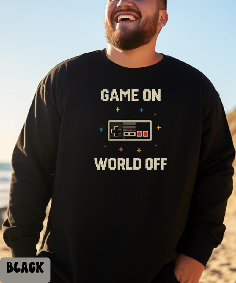 May include: Black sweatshirt with the text "GAME ON WORLD OFF" in pixelated white letters. A pixelated controller graphic is centered between the text. The sweatshirt is a crew neck style.