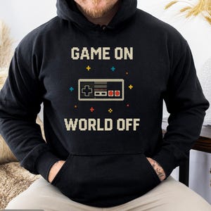 May include: Black hoodie with the text "GAME ON WORLD OFF" in pixelated font. A pixelated controller graphic is centered below the text. The hoodie has a front pocket and a drawstring hood.