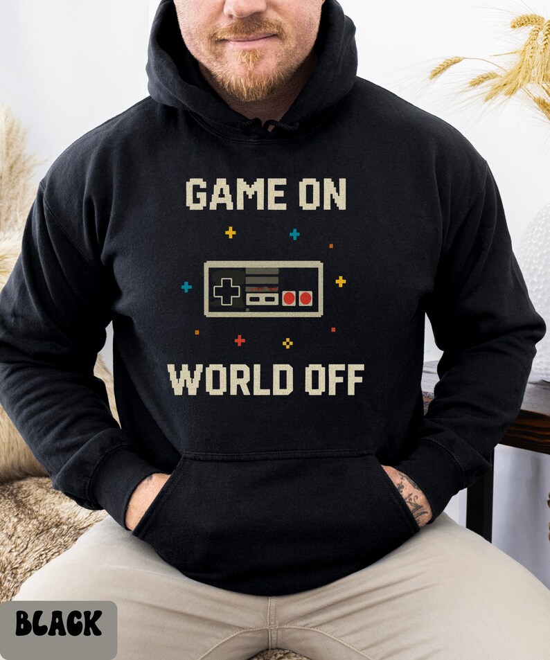May include: Black hoodie with the text "GAME ON WORLD OFF" in pixelated font. A pixelated controller graphic is centered below the text. The hoodie has a front pocket and a drawstring hood.