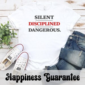 May include: White t-shirt with the words "SILENT DISCIPLINED DANGEROUS." in black and red. The shirt is paired with blue jeans and white canvas sneakers. The text "Happiness Guarantee" is also visible.