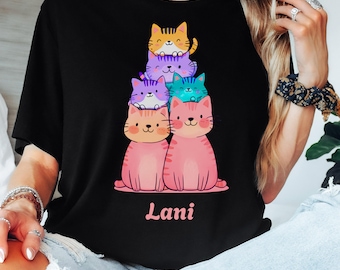 May include: Black t-shirt featuring a stack of cartoon cats in various colors: orange, purple, blue, and pink. The name "Lani" is printed in pink script below the cats. The word "BLACK" is printed in a gray rectangle at the bottom.