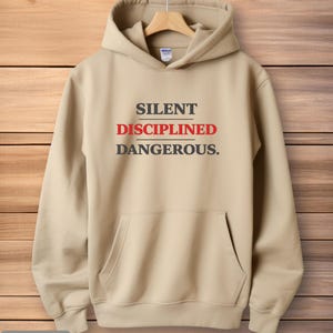 May include: Sand-colored hoodie with a front pocket and a drawstring hood. The text "SILENT DISCIPLINED DANGEROUS." is printed in black and red across the chest. The hoodie is displayed on a wooden hanger.