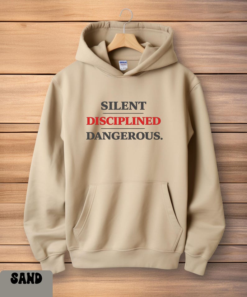 May include: Sand-colored hoodie with a front pocket and a drawstring hood. The text "SILENT DISCIPLINED DANGEROUS." is printed in black and red across the chest. The hoodie is displayed on a wooden hanger.