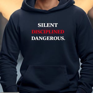 May include: A navy blue hoodie with a front pocket and the words "SILENT DISCIPLINED DANGEROUS." in white and red lettering. The word "BLACK" is at the bottom left corner of the image.