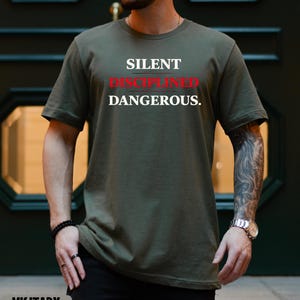 May include: Military green t-shirt with the words "SILENT DISCIPLINED DANGEROUS" in white and red lettering. The shirt is a solid color and has short sleeves. The shirt is made of a soft, comfortable material.