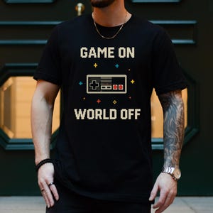 May include: Black t-shirt with the text "GAME ON WORLD OFF" in pixelated white letters. A pixelated video game controller graphic is centered between the text. The word "BLACK" is printed at the bottom.