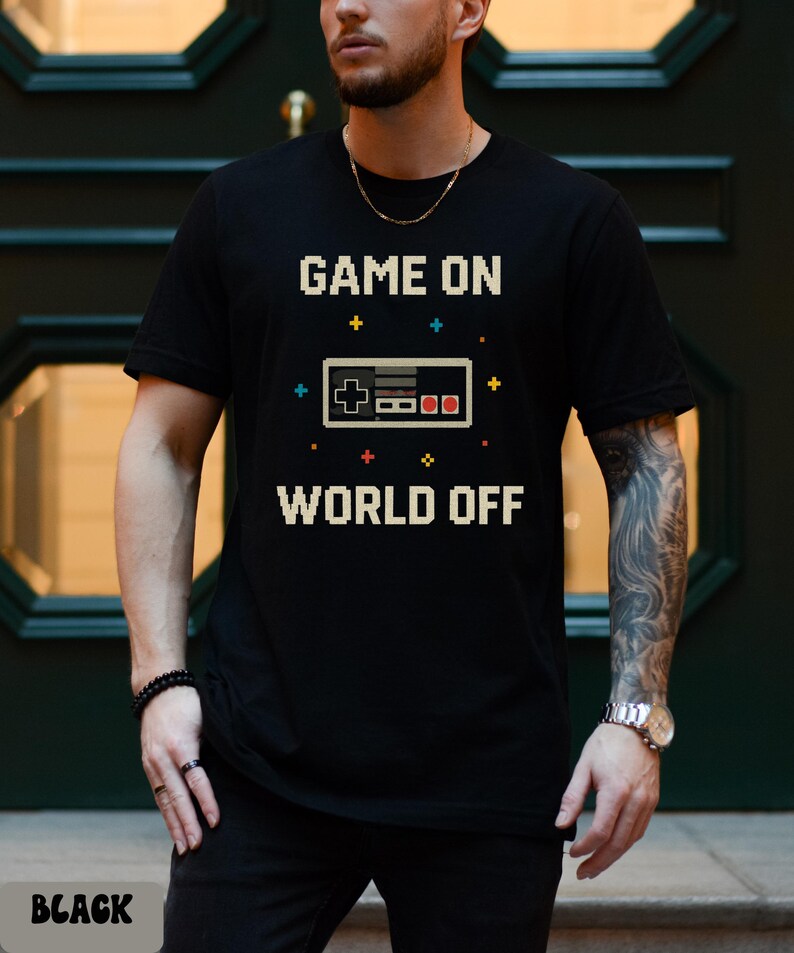 May include: Black t-shirt with the text "GAME ON WORLD OFF" in pixelated white letters. A pixelated video game controller graphic is centered between the text. The word "BLACK" is printed at the bottom.