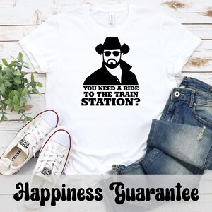 K&ouml;nnte beinhalten: Wei&szlig;es T-Shirt mit einer schwarzen Grafik eines Mannes mit Cowboyhut und Sonnenbrille, mit dem Text "YOU NEED A RIDE TO THE TRAIN STATION?". Das Shirt wird mit blauen Jeans und wei&szlig;en Turnschuhen gezeigt.