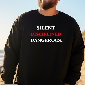 May include: Black crew neck sweatshirt with the words "SILENT DISCIPLINED DANGEROUS." in white and red lettering. The word "BLACK" is in a gray box at the bottom of the image. The sweatshirt is a solid black color.