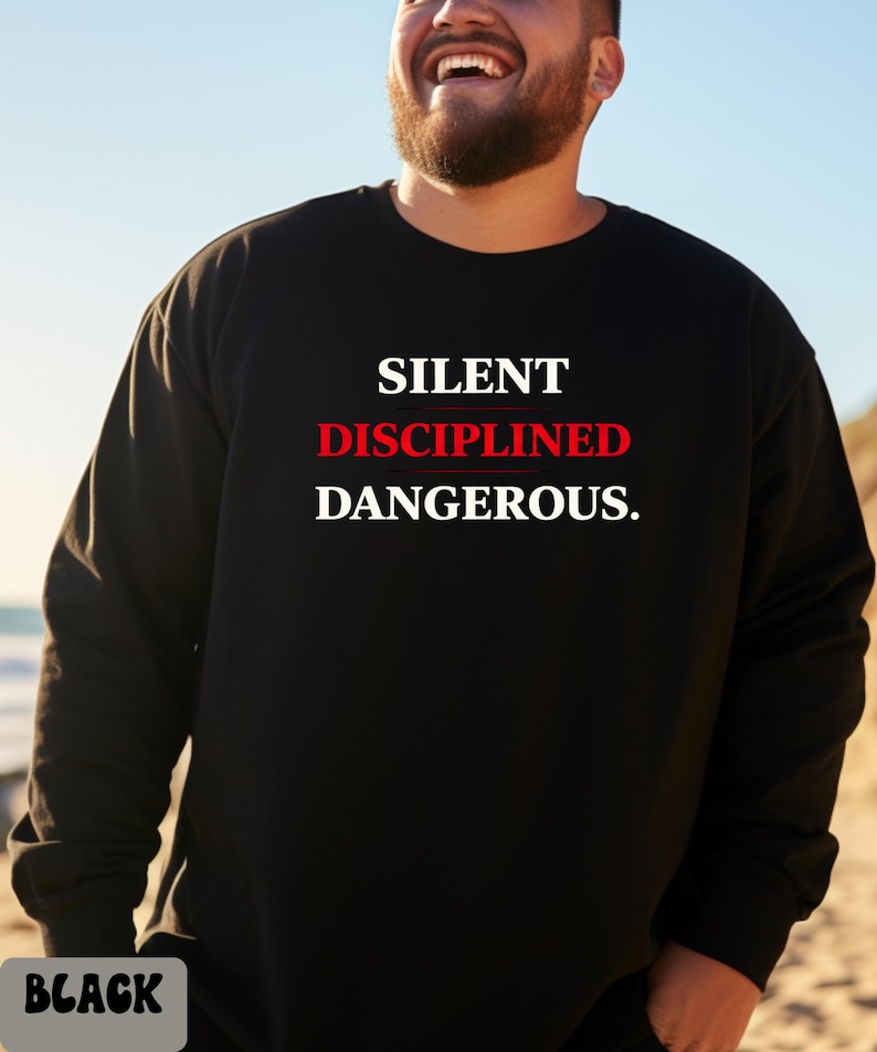 May include: Black crew neck sweatshirt with the words "SILENT DISCIPLINED DANGEROUS." in white and red lettering. The word "BLACK" is in a gray box at the bottom of the image. The sweatshirt is a solid black color.