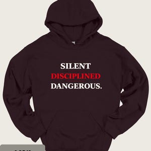 May include: Dark chocolate-colored hoodie with a front pocket and hood. The text "SILENT DISCIPLINED DANGEROUS" is printed in white and red. The bottom left corner of the image has the text "DARK CHOCOLATE".