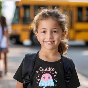 Puede incluir: Camiseta negra con un diseño de mariquita rosa de dibujos animados, una manta azul y rosa, y las palabras "Cuddle Bug" y "Name". Una niña con una mochila frente a un autobús escolar amarillo en el fondo.
