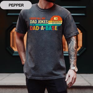 May include: Dark gray t-shirt with the text "I KEEP ALL MY DAD JOKES IN A DAD-A-BASE" in a retro design. The graphic includes a floppy disk icon in orange, yellow, and blue.