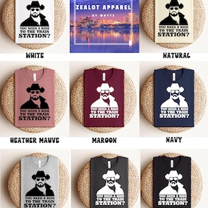 K&ouml;nnte beinhalten: Eine Auswahl an T-Shirts in verschiedenen Farben, jeweils mit einer Grafik eines Mannes mit Cowboyhut und Sonnenbrille sowie dem Text "YOU NEED A RIDE TO THE TRAIN STATION?". Farben sind Wei&szlig;, Natur, Heather Mauve, Maroon, Navy, Athletic Heather, Dark Grey Heather und Schwarz.