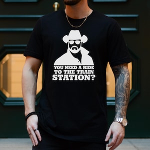 K&ouml;nnte beinhalten: Schwarzes T-Shirt mit wei&szlig;em Aufdruck eines Cowboys mit Sonnenbrille und Hut. Darunter steht der Text "YOU NEED A RIDE TO THE TRAIN STATION?". Das Wort "BLACK" ist unten aufgedruckt.