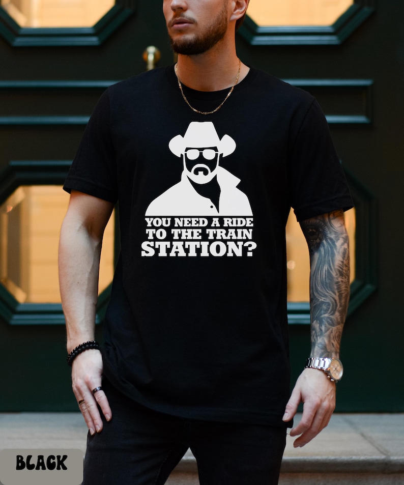 K&ouml;nnte beinhalten: Schwarzes T-Shirt mit wei&szlig;em Aufdruck eines Cowboys mit Sonnenbrille und Hut. Darunter steht der Text "YOU NEED A RIDE TO THE TRAIN STATION?". Das Wort "BLACK" ist unten aufgedruckt.