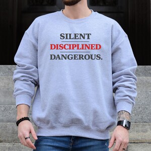 May include: A light gray sweatshirt with the words "SILENT DISCIPLINED DANGEROUS." printed in black and red. The sweatshirt is a sport grey color.