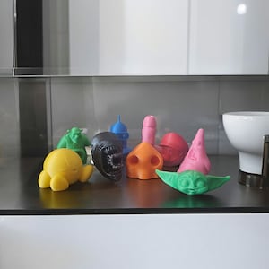 May include: A collection of 10 small, colourful, 3D printed figurines. The figurines include a green Shrek character, a yellow character with a round head, a blue alien character, a pink character with a star shape, a red character with a round head, a green character with a round head, a black character with a skull-like head, a pink character with a round head, a brown character with a round head, and a pink character with a round head.