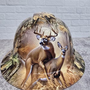 May include: A hard hat with a detailed graphic of two deer in a forest setting. The hat features a camouflage pattern on the brim and sides, with the deer prominently displayed on the crown.